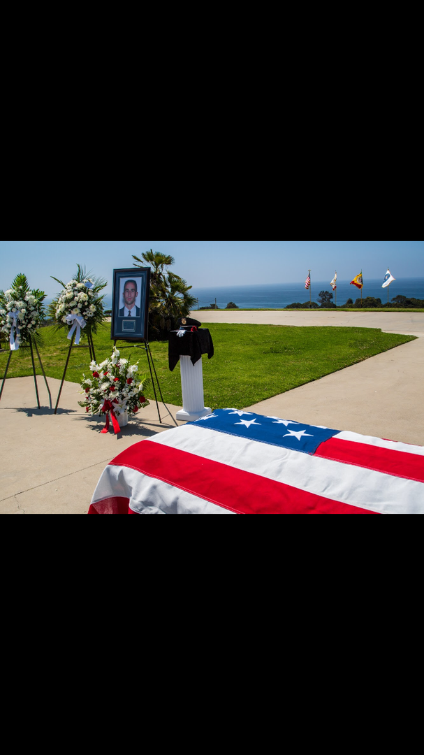 A casket draped in an American flag sits on a paved area with flower arrangements and a framed portrait near the ocean.