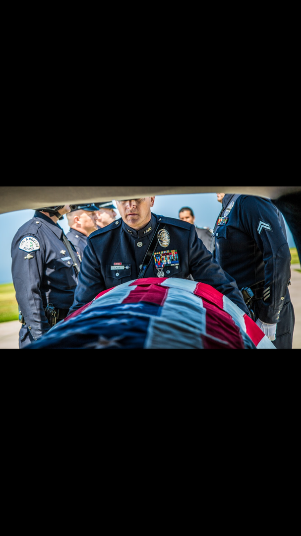 Police officers in formal uniforms solemnly carry a casket draped in an American flag.