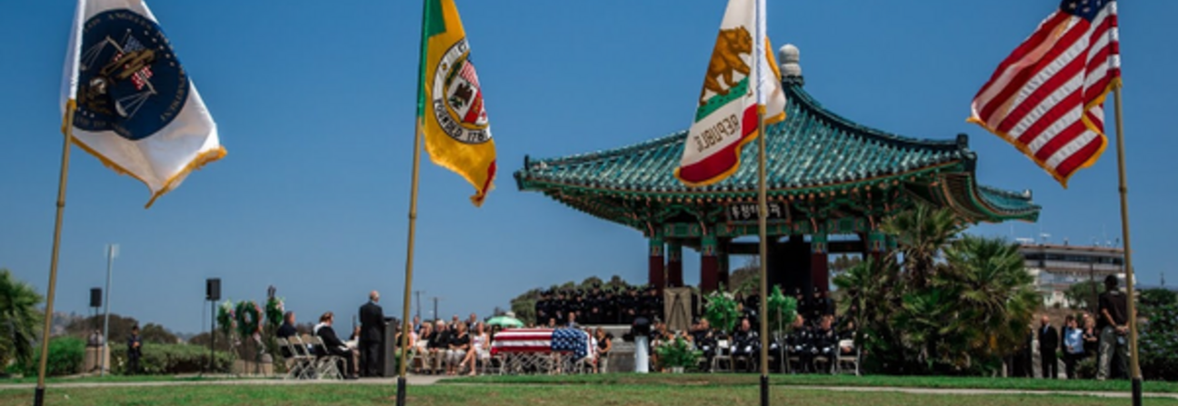 Four flags fly in front of a traditional Korean-style pavilion during an outdoor ceremony under a clear blue sky.