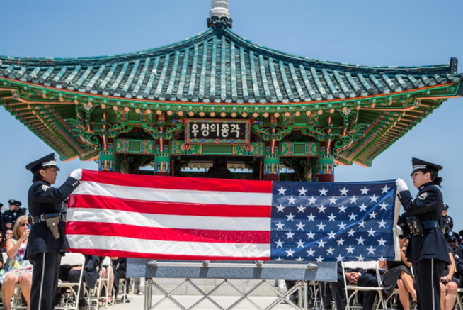 Two uniformed officers hold a large American flag in front of a traditional Korean pavilion.