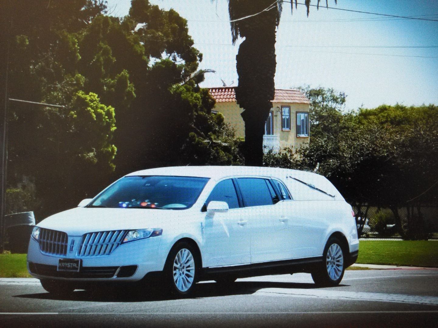 A white Lincoln MKT hearse driving on a sunny street near trees and a house.