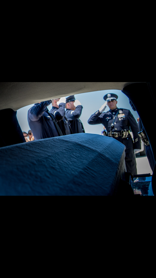 Police officers in formal uniform stand in a line and salute as a casket is transported.