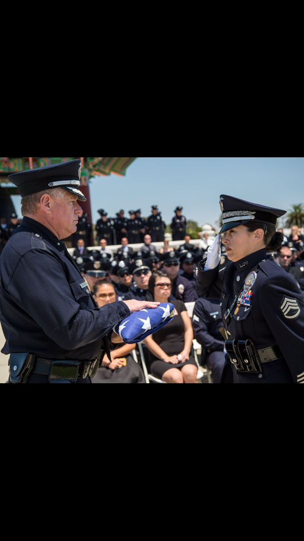 A uniformed officer presents a folded American flag to a uniformed colleague during an outdoor memorial service.