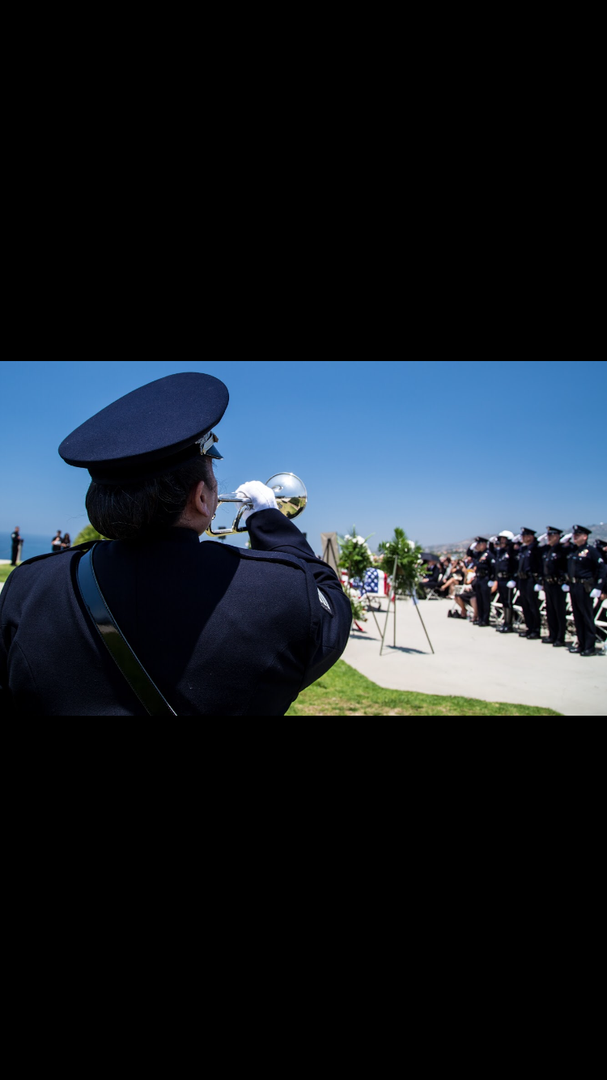 A uniformed officer plays a trumpet during an outdoor military-style memorial ceremony with other personnel in formation.