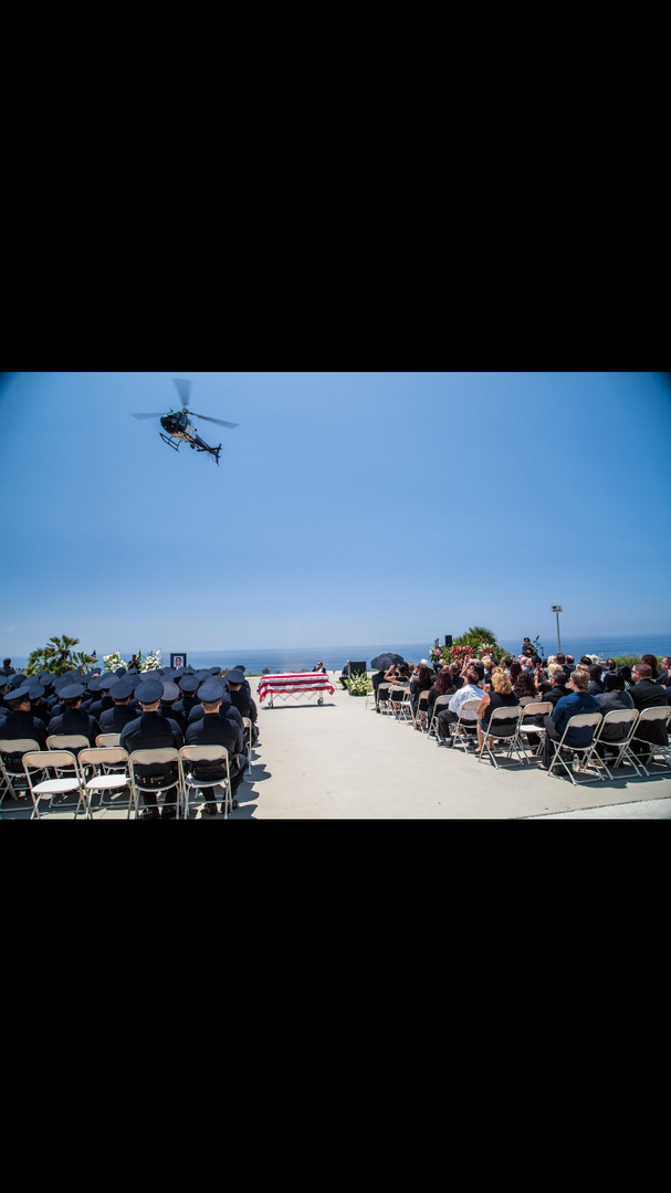 A helicopter flies over an outdoor memorial service with seated attendees facing a central American flag.