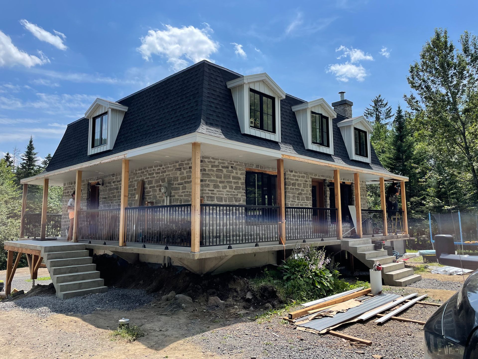 Une maison en pierre avec toit mansard&eacute; noir, porche enveloppant avec colonnes en bois et rampes m&eacute;talliques, en construction