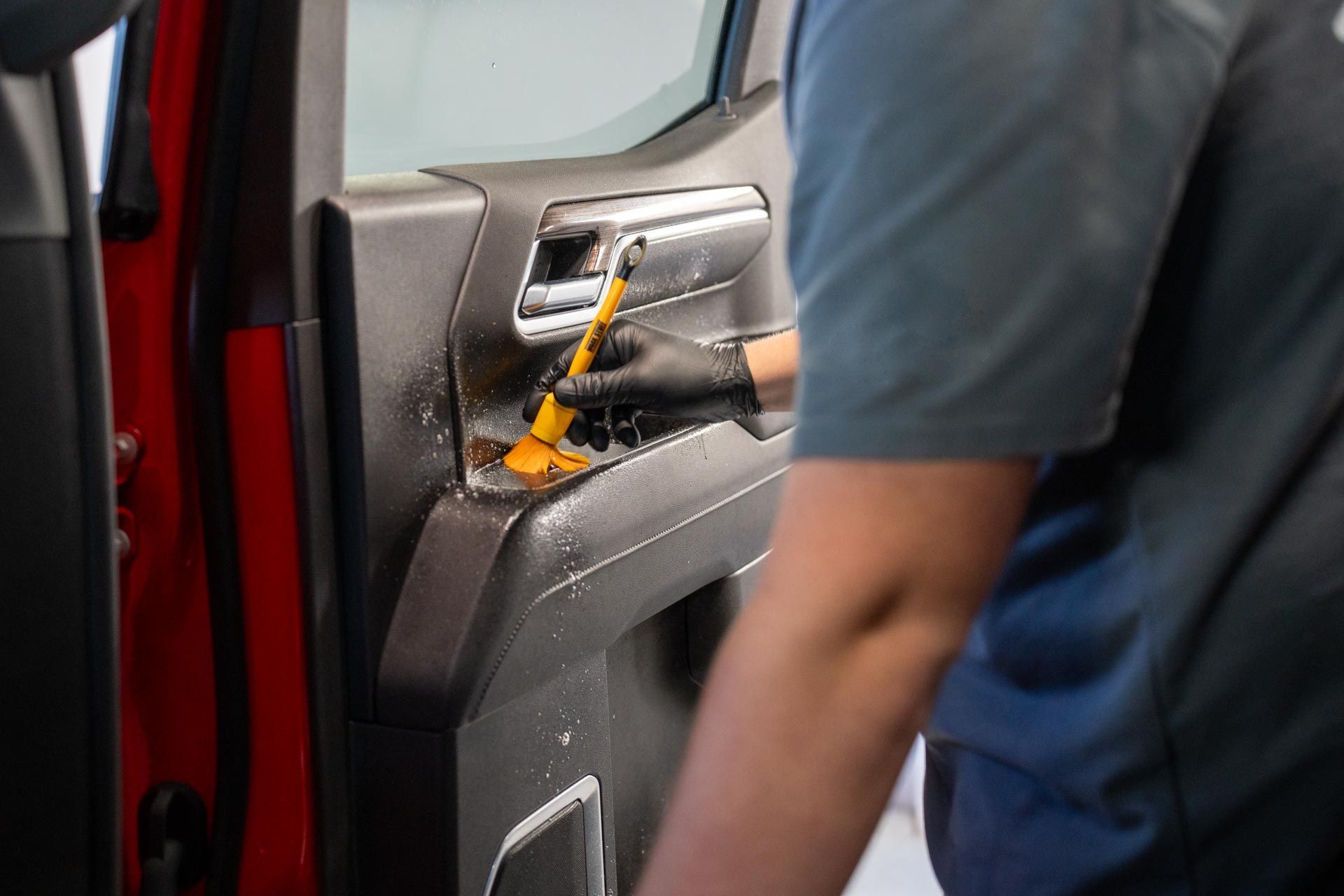 Person cleaning a car door panel with a brush, wearing gloves, in a vehicle's interior.