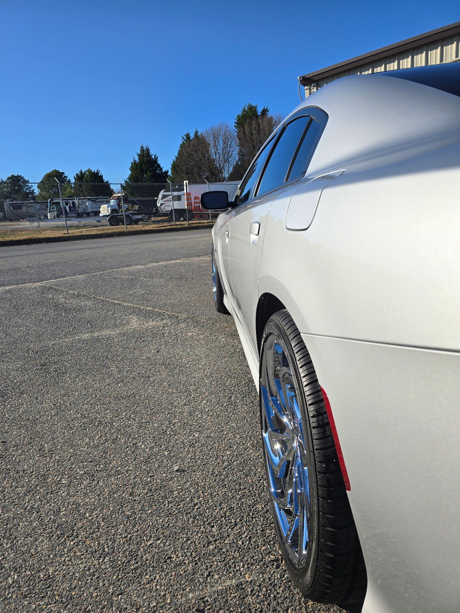 A silver car parked on a gravel lot on a sunny day, focusing on the rear passenger side and chrome wheel.