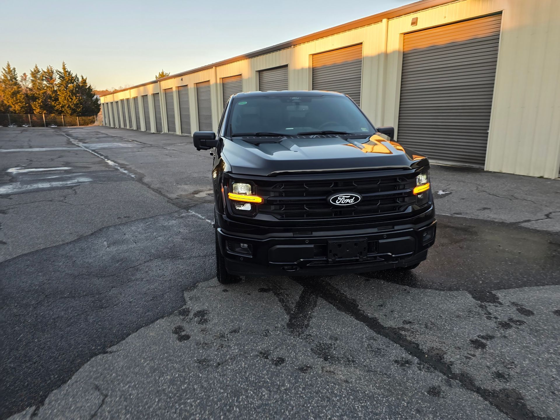 A black Ford F-150 truck parked on an asphalt lot in front of several storage units during a sunset.