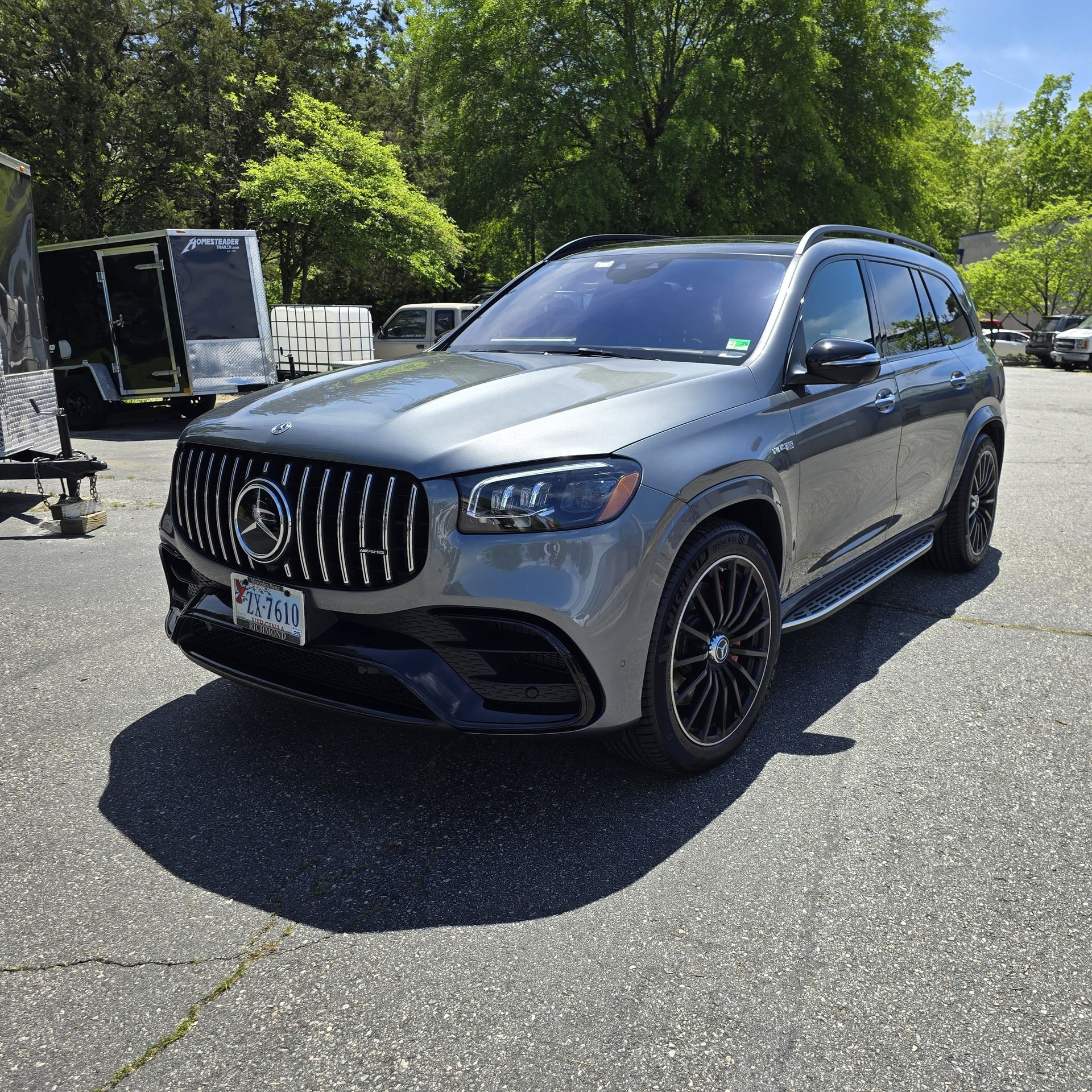 Gray Mercedes-Benz GLS SUV parked on pavement. Black grill, wheels. Trailer and trees in background.