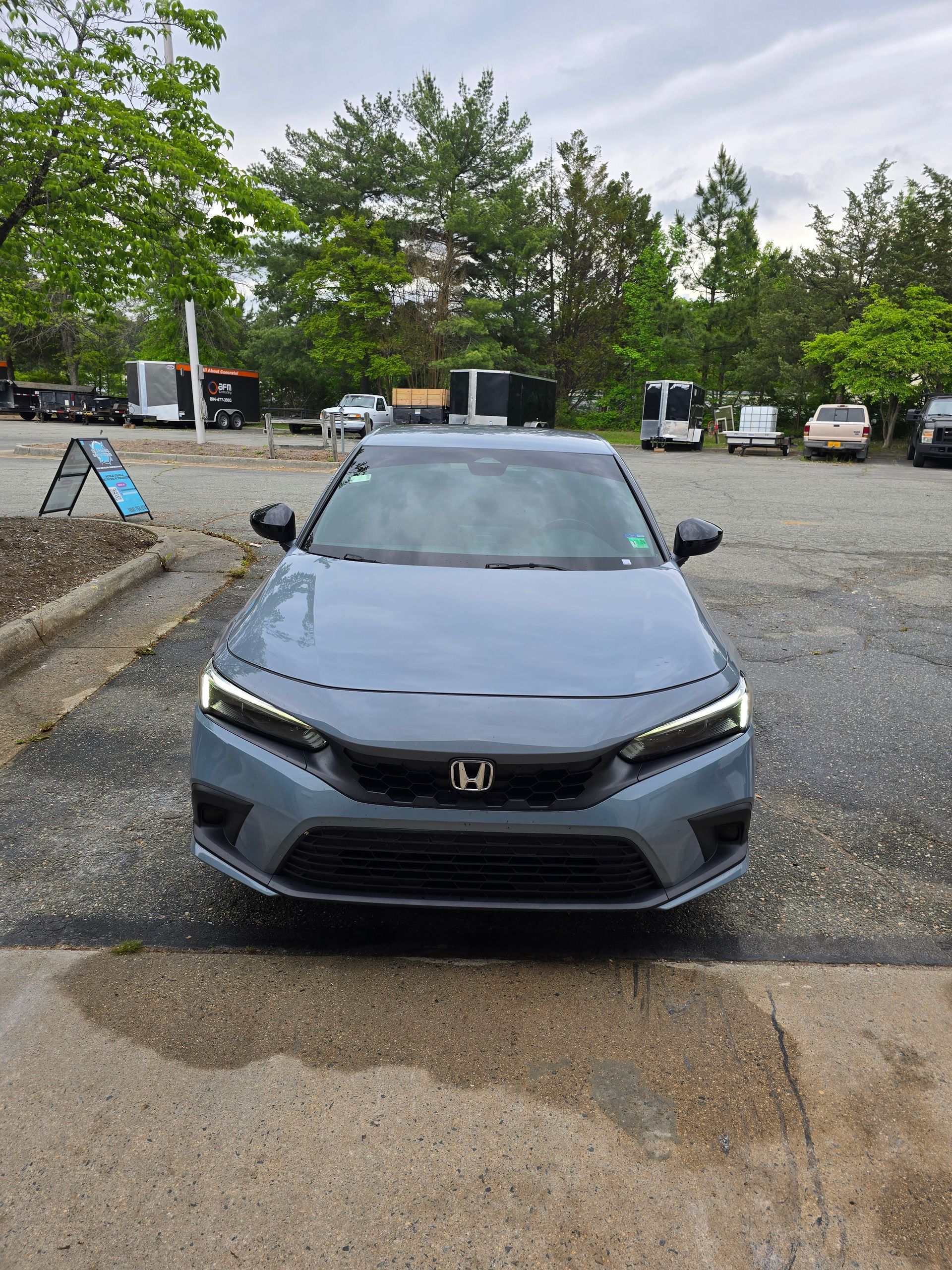 A blue-gray Honda Civic parked outdoors on a wet paved surface with other vehicles and trees in the background.