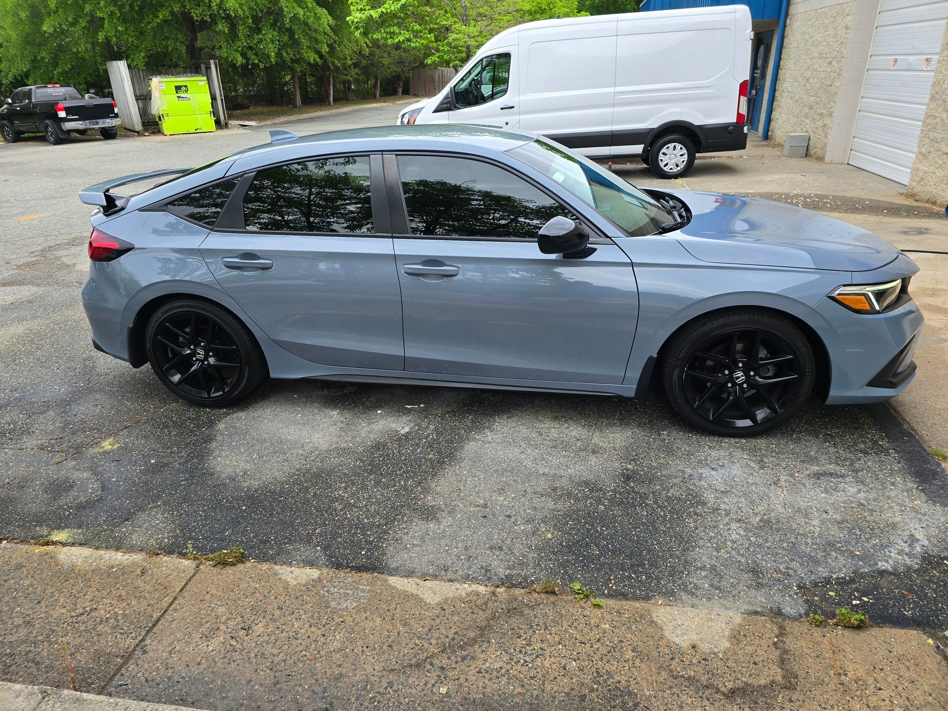 Gray Honda Civic with black wheels parked on pavement, white van in the background.
