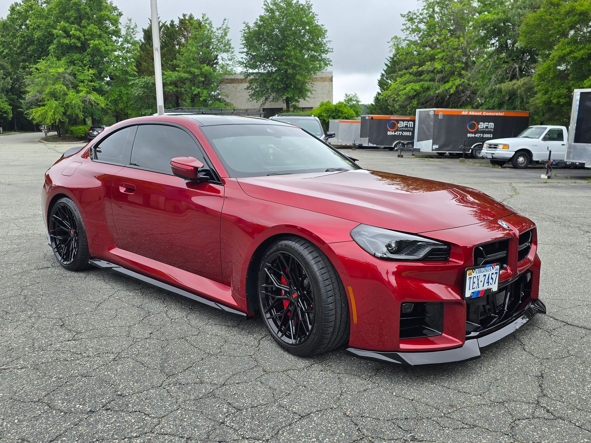 A shiny, red BMW M2 coupe parked on an asphalt lot with black wheels and a front splitter, seen from a side angle.