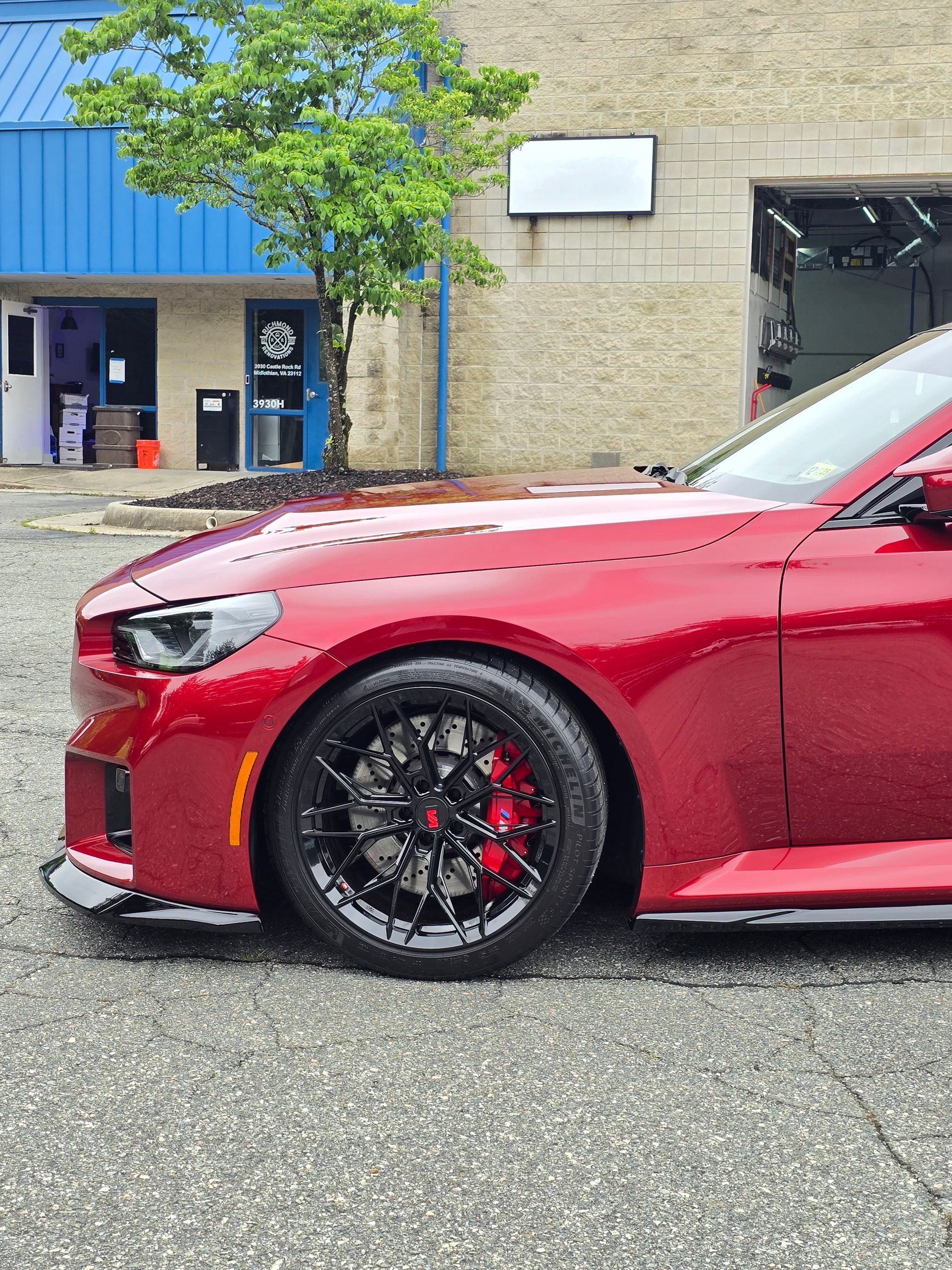 A close-up of a glossy red sports car parked on an asphalt lot in front of a building with a tree.