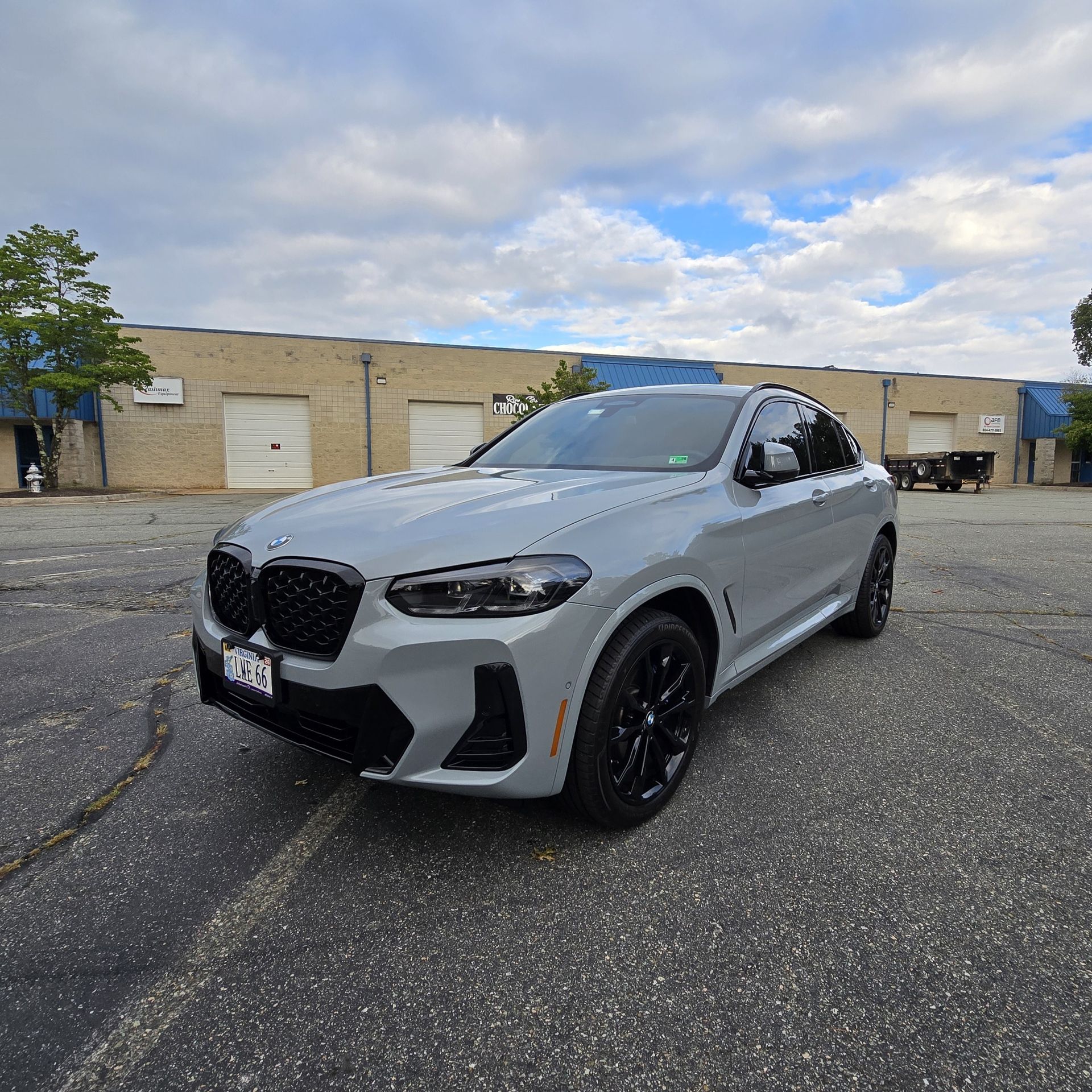 Gray BMW X4 SUV with black wheels parked on asphalt in front of a building with a cloudy sky overhead.