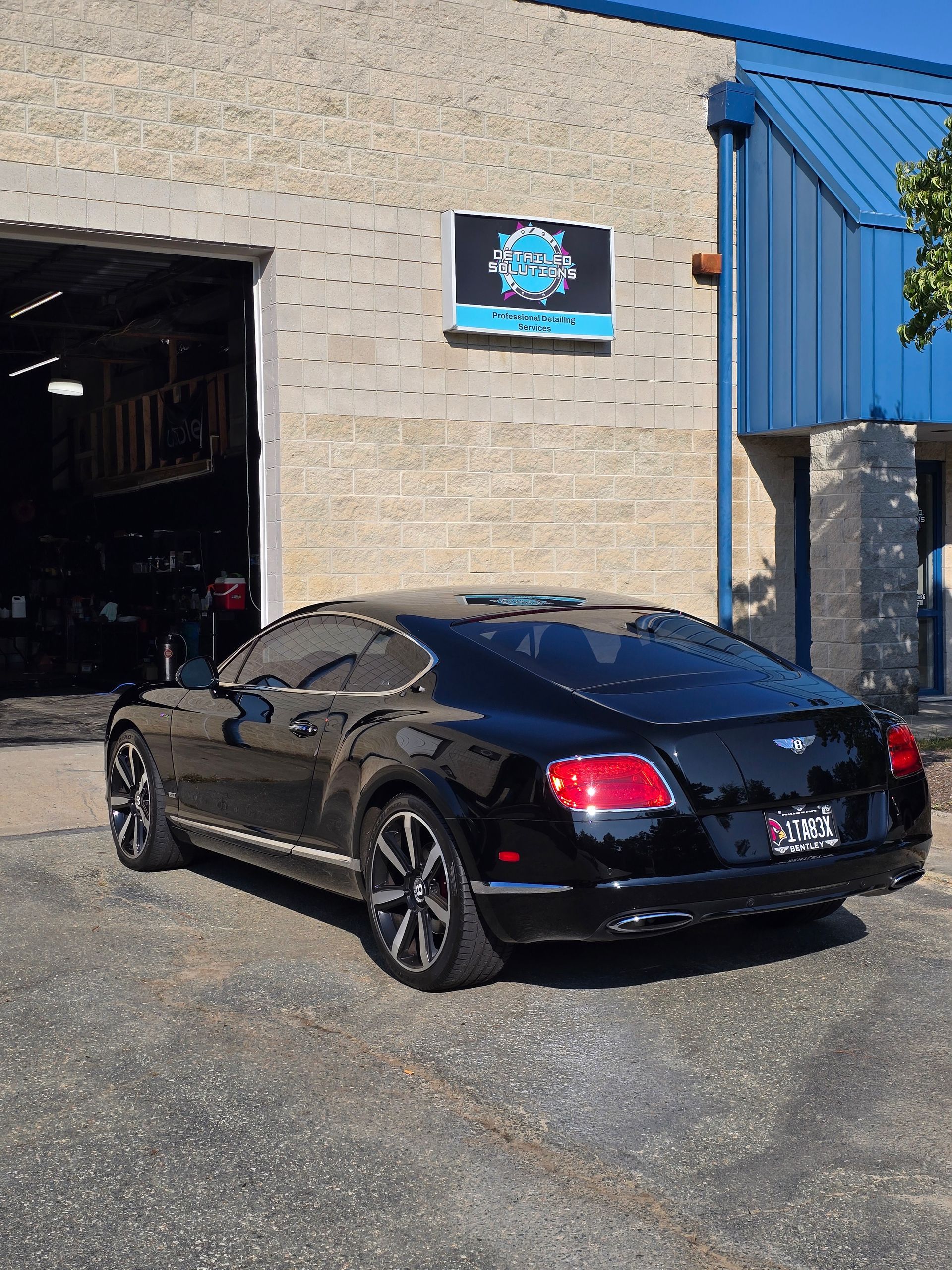 Black Bentley parked outside a blue-trimmed auto shop with a sign above the door.