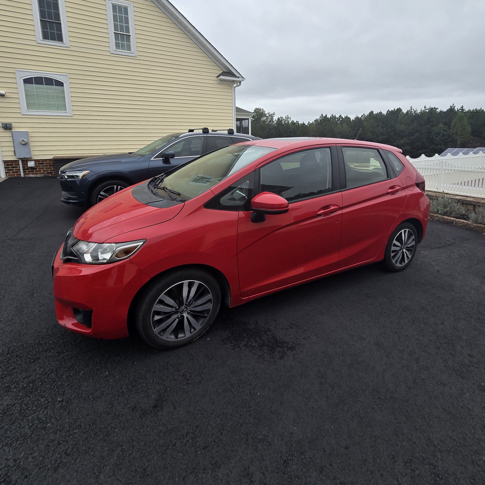 Red Honda Fit parked on black asphalt driveway. A dark blue car is in the background. Beige house. Overcast sky.