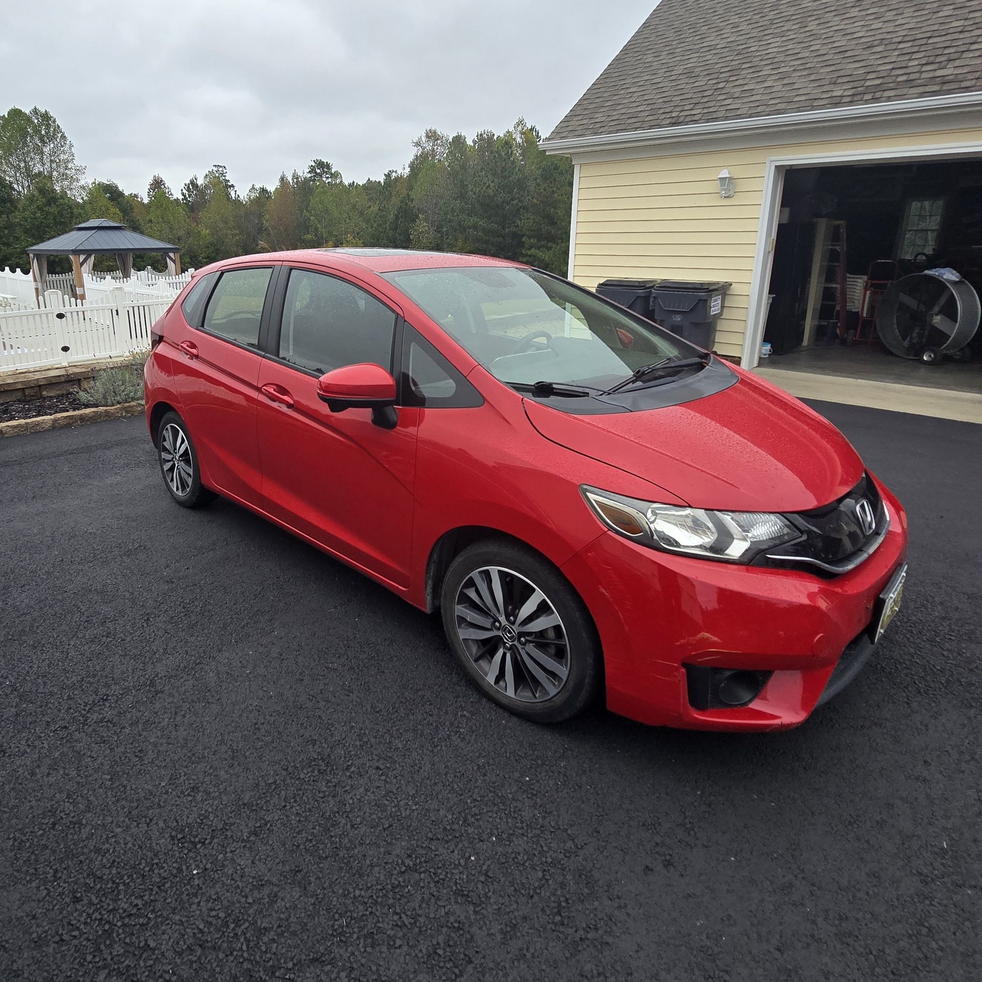 Red Honda Fit parked on a black asphalt driveway, facing forward. New Jersey license plate. Cloudy sky in background.