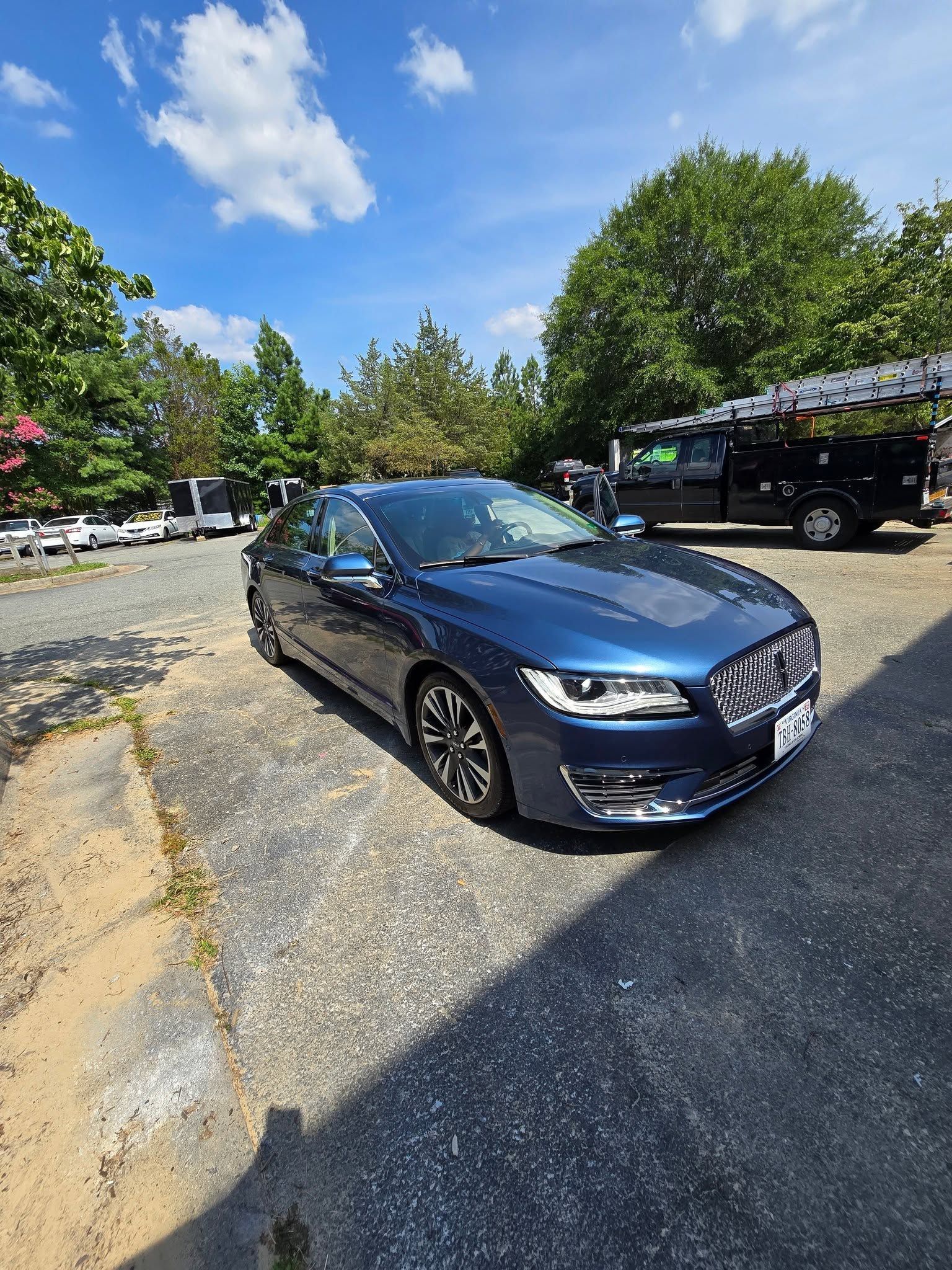A dark blue ceramic-coated sedan parked on a gravel lot under a bright blue sky.