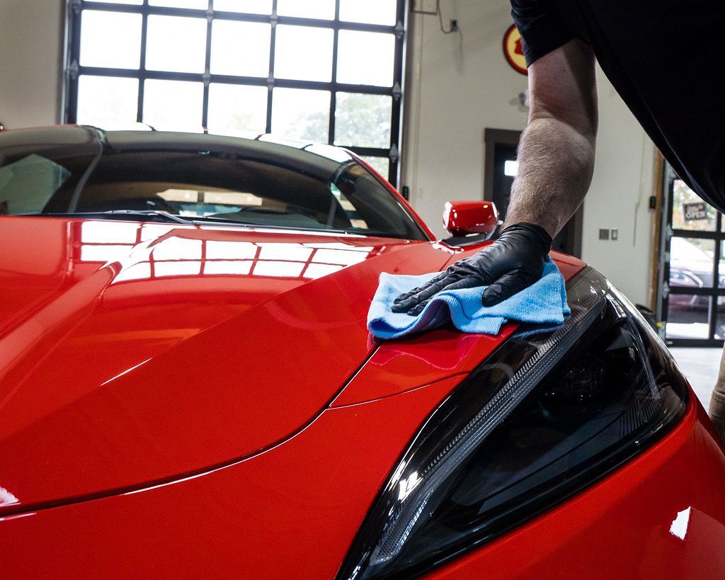 Person wiping a ceramic coated red sports car’s front headlight with a blue cloth in a garage