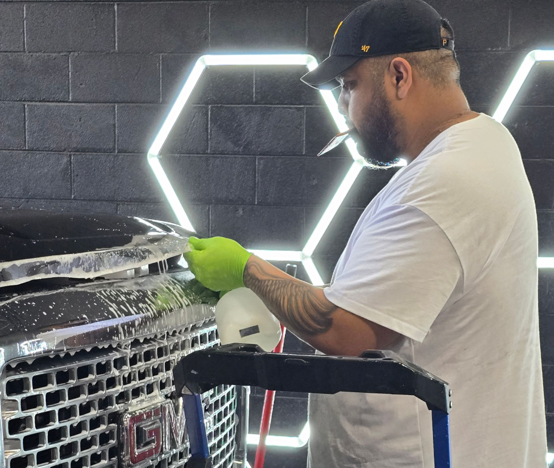 Man washing a black GMC truck. He wears a cap, gloves and white shirt, using a foam sprayer. Hexagon lights in the background.