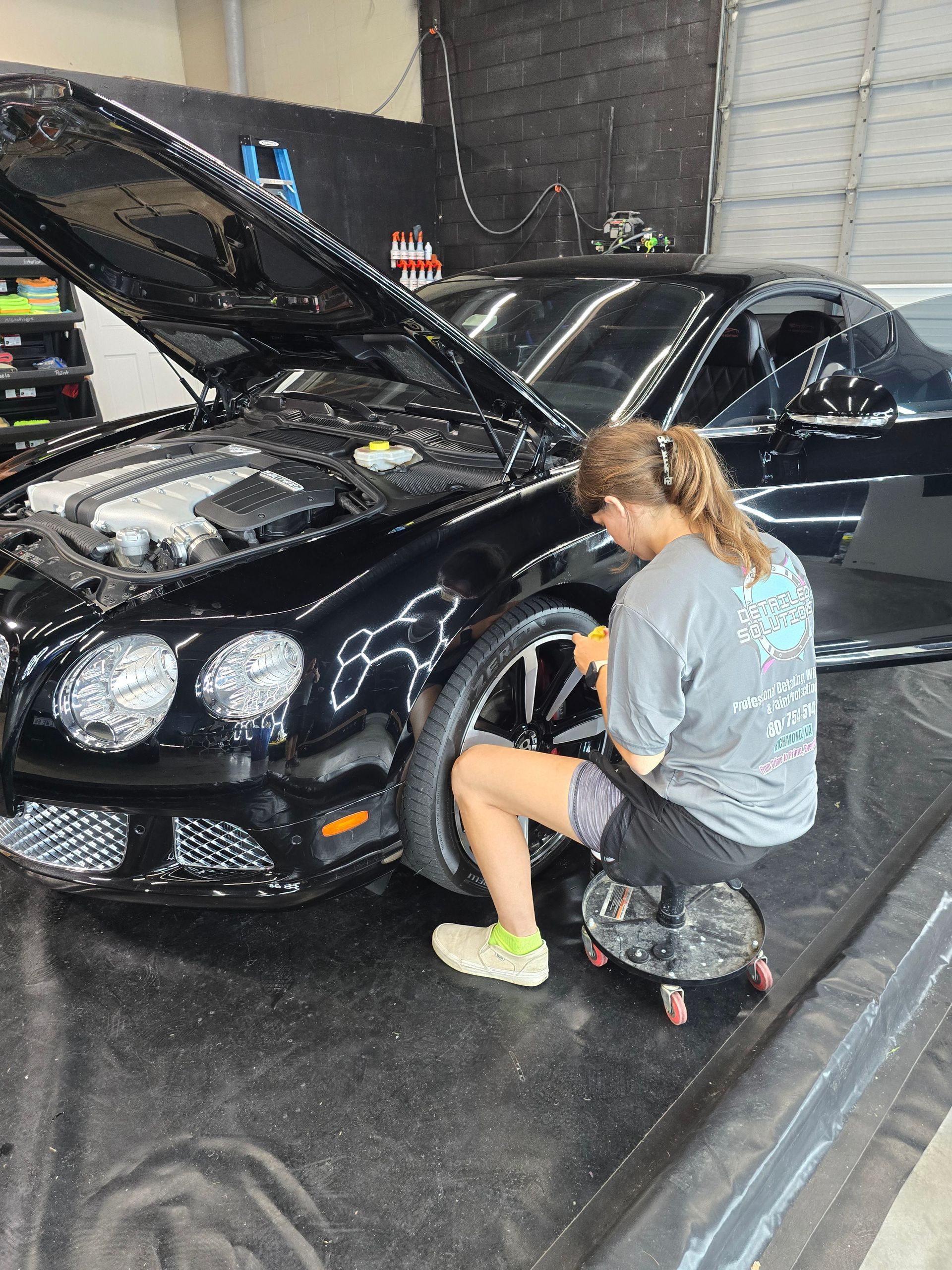 Woman kneeling beside a black sports car with the hood open in a garage applying PPF.