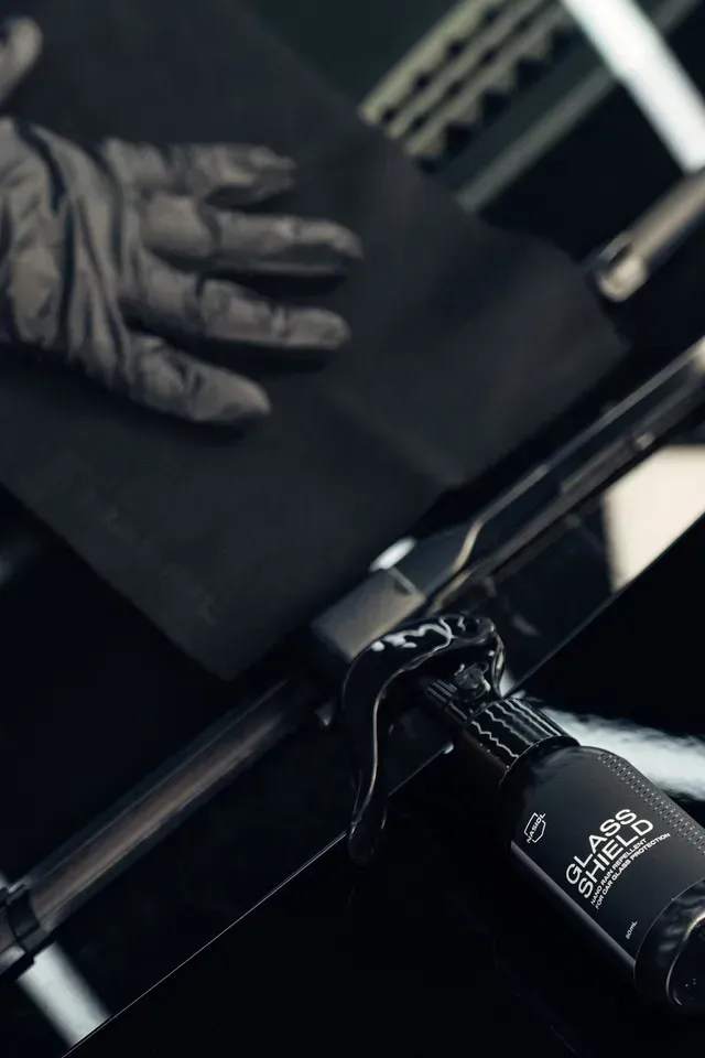 A gloved hand wipes a dark surface near a bottle of Glass Shield professional coating product.