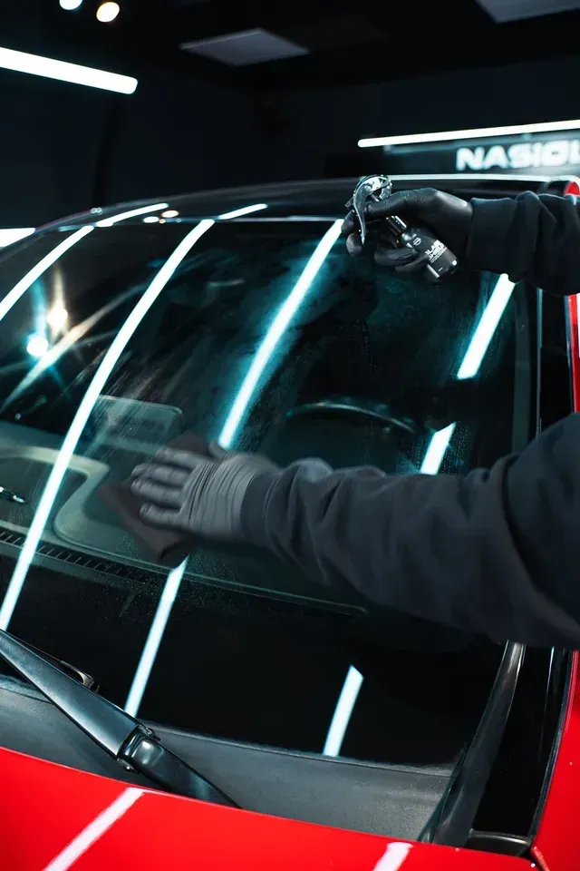 A person in black gloves cleans a car's windshield with a spray bottle and a microfiber cloth in a brightly lit garage.