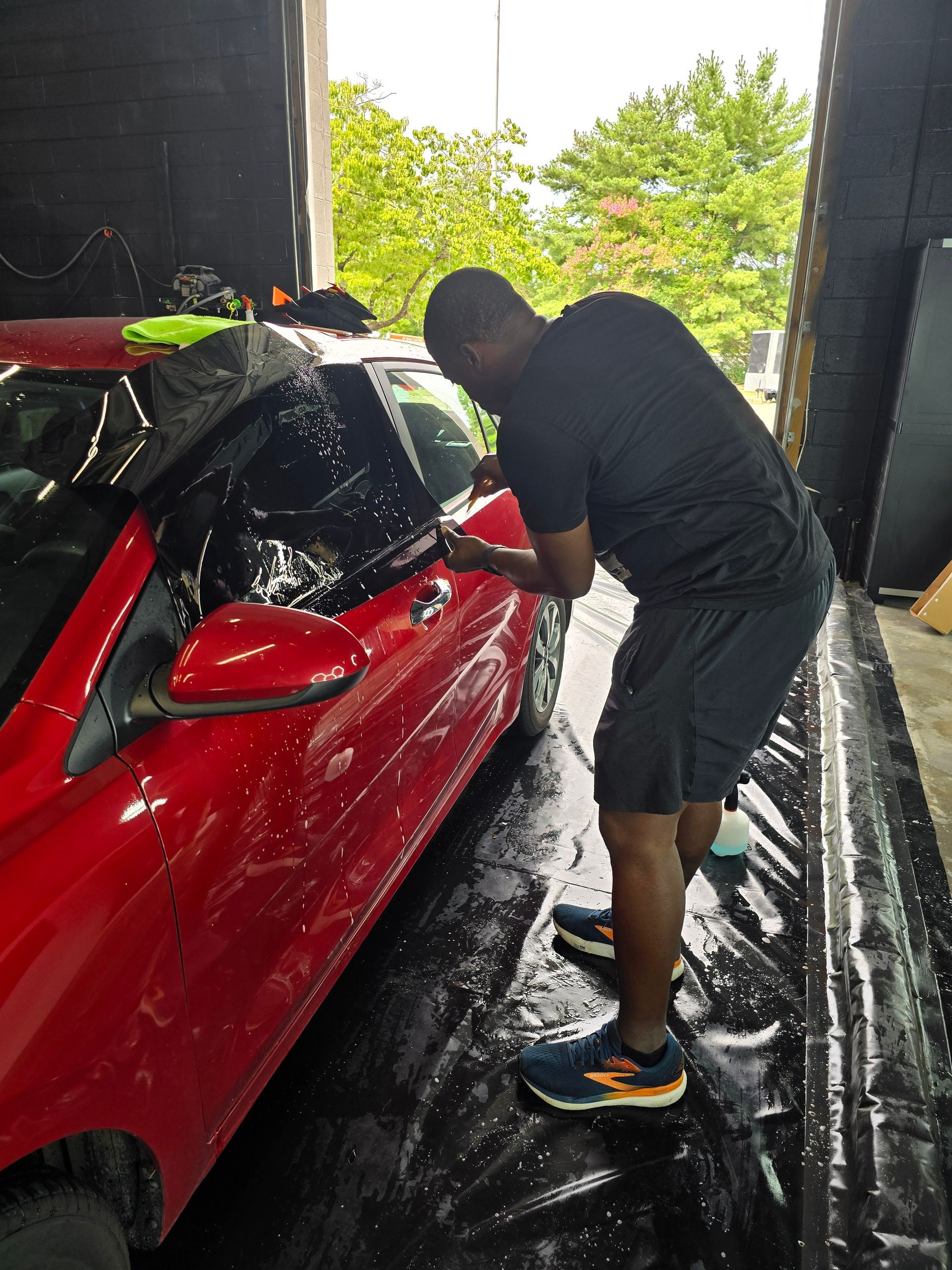 Technician professionally installing window tint on a red car inside an automotive detailing shop