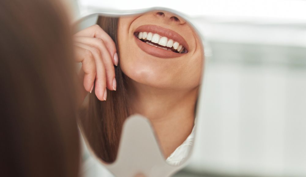 A close-up of a person smiling, reflected in a small handheld mirror, focusing on their teeth and lower face.