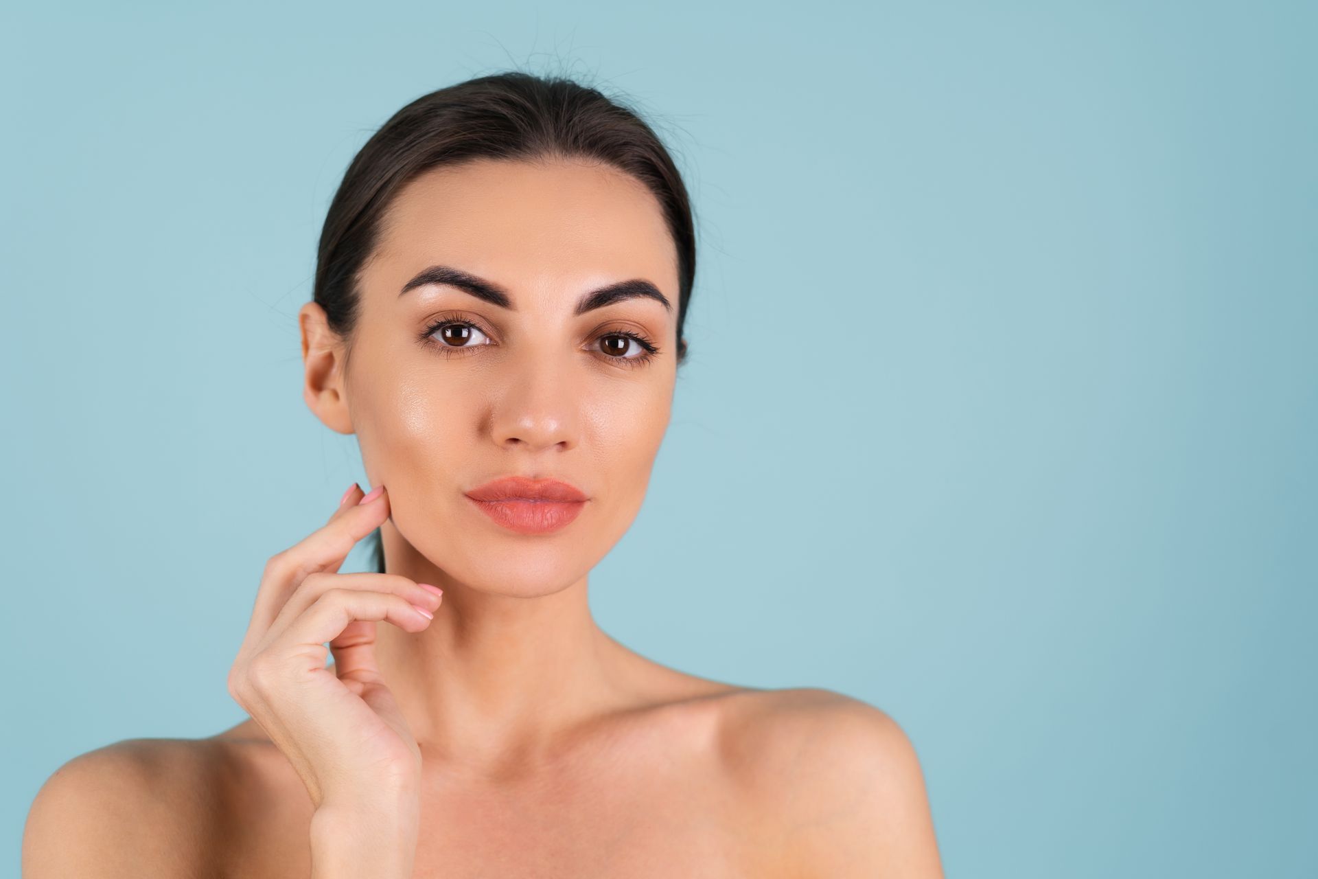 Portrait of a woman with bare shoulders touching her cheek against a light blue background