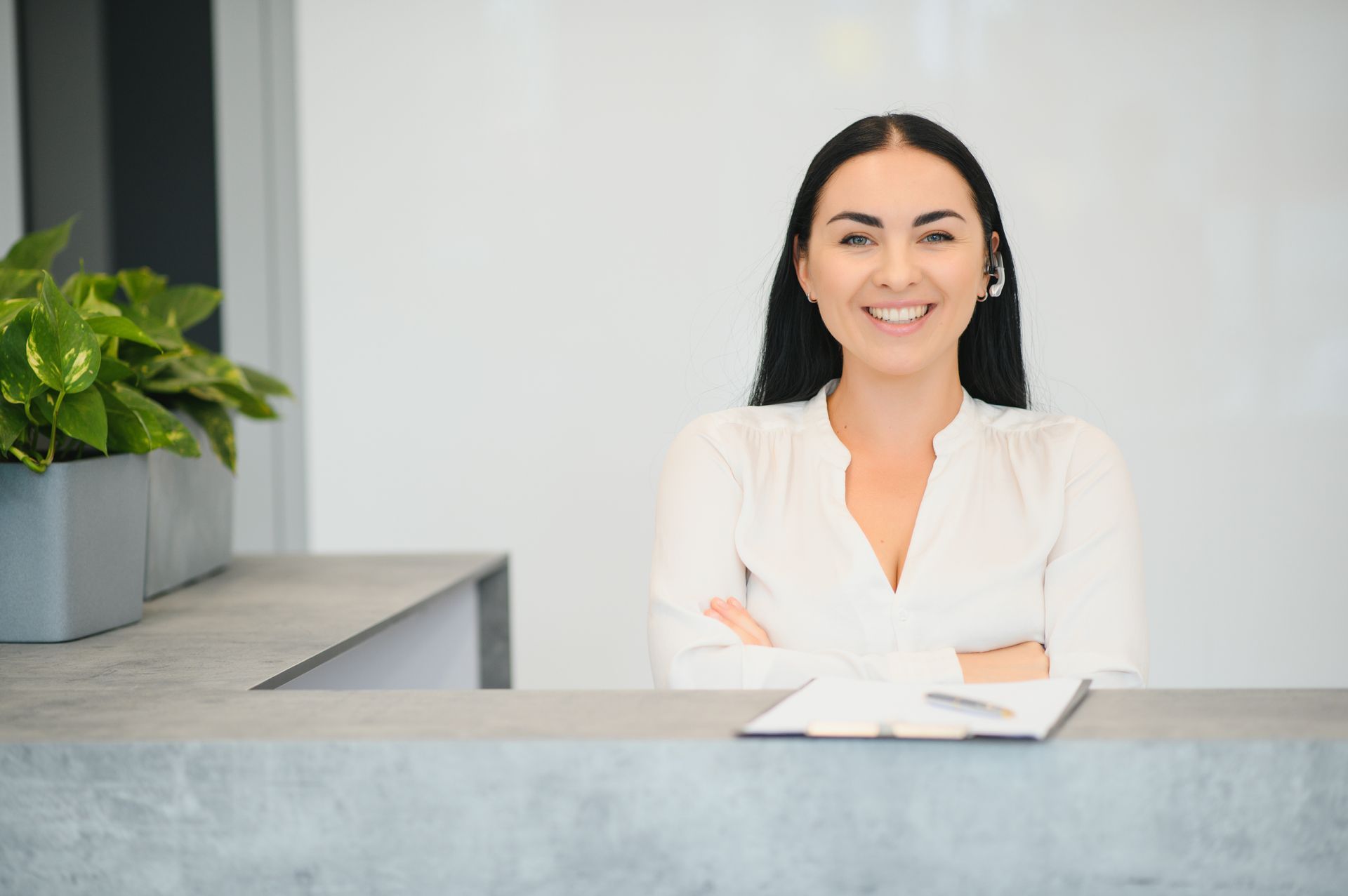 Woman with a smile, standing behind a gray reception desk. She has dark hair, arms crossed, and wears a white shirt. Plant on the left.