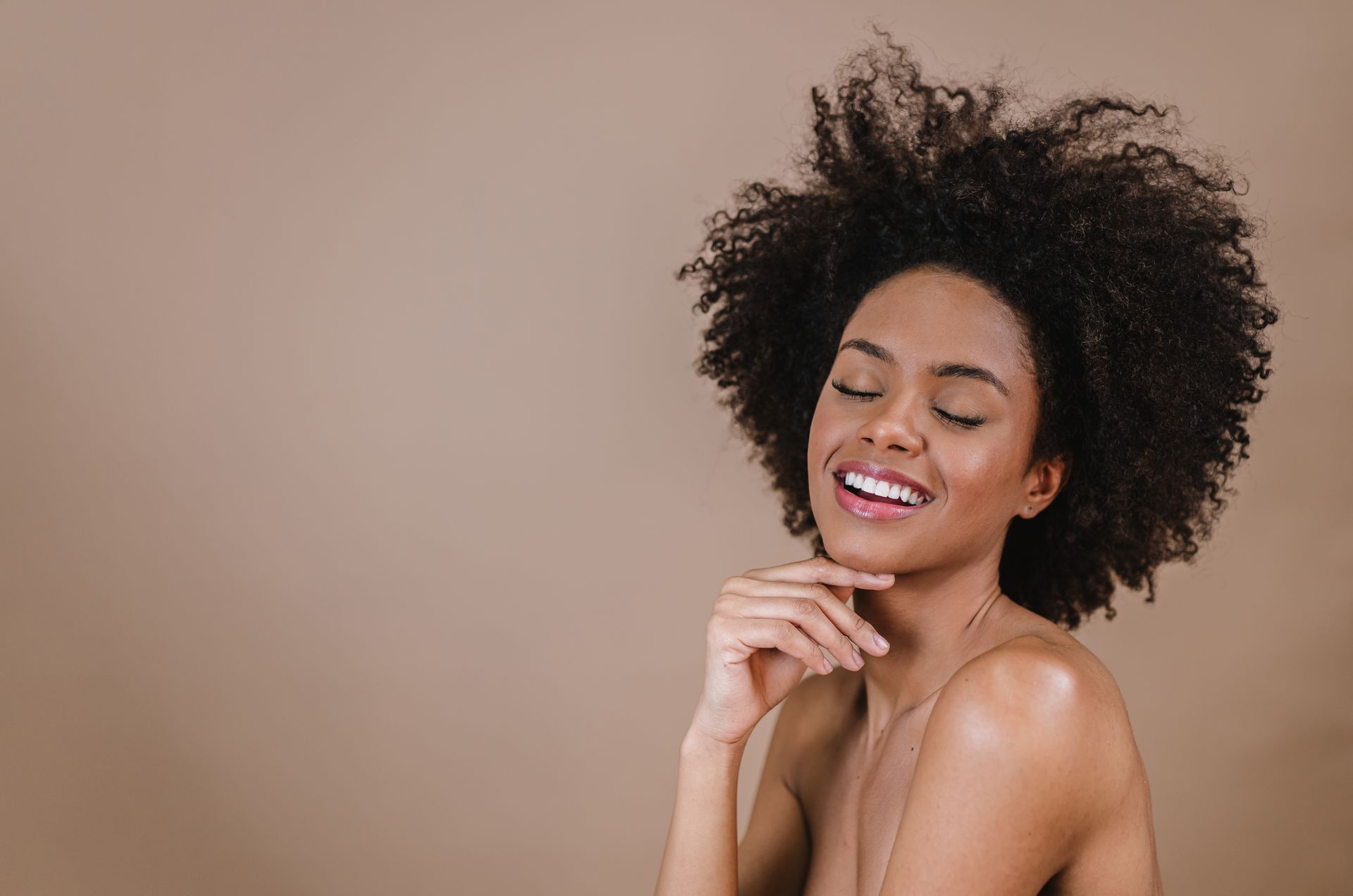 Smiling woman with curly hair posing against a beige background, resting her chin on her hands