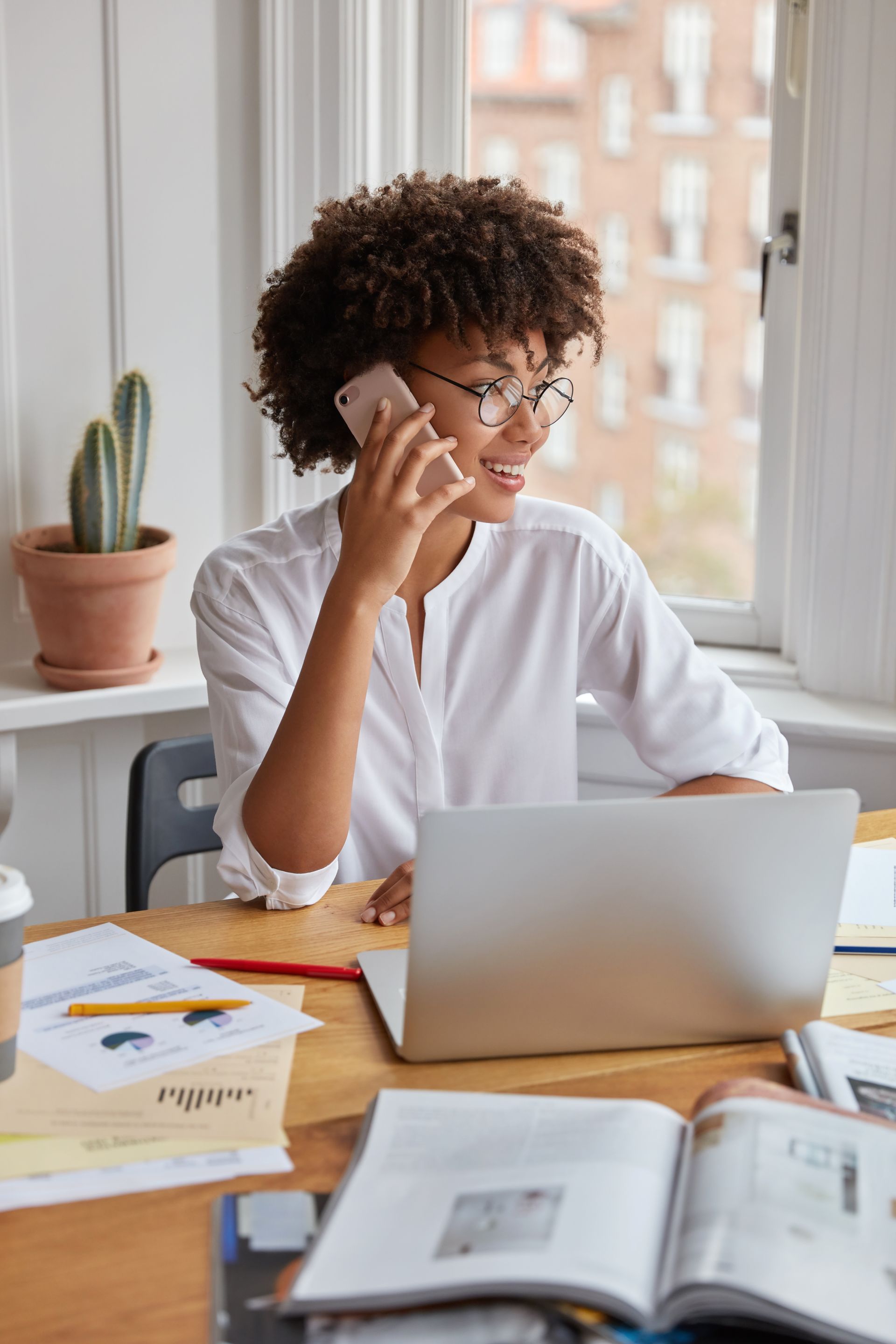 Woman with glasses, curly hair, and white shirt smiles while talking on her phone at a desk with a laptop, in front of a window.