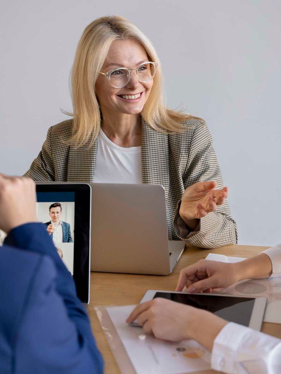 Blonde woman with glasses smiles, gesturing during a meeting at a table with a laptop, tablet, and person on video call.