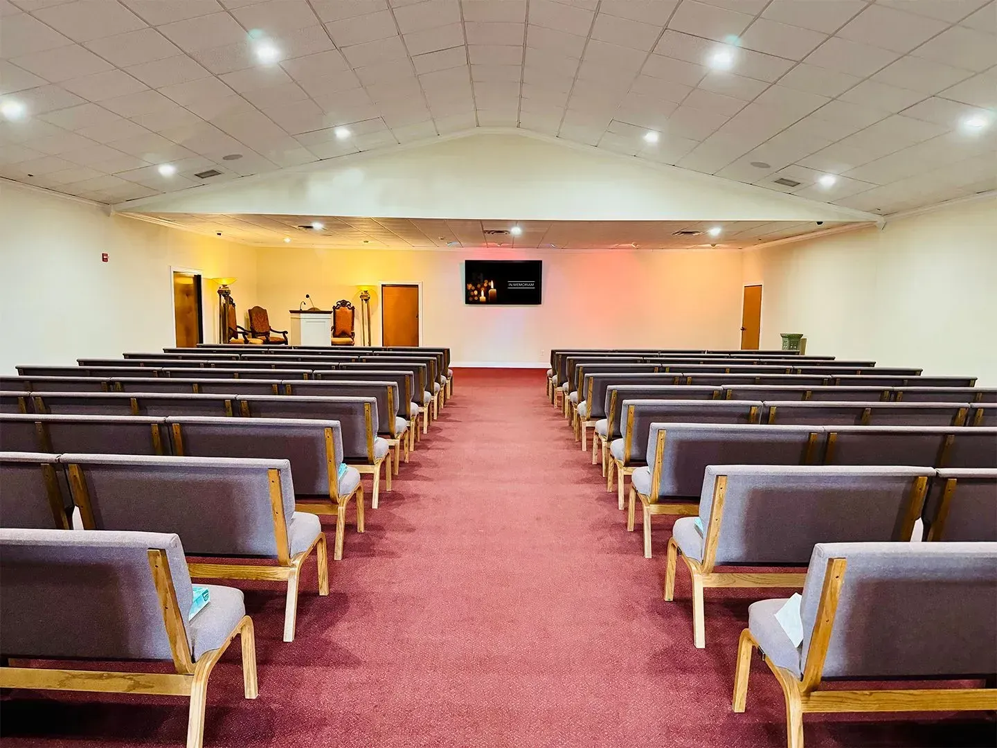 An empty chapel with rows of grey padded wooden pews facing a central stage area with a screen and red carpet flooring.