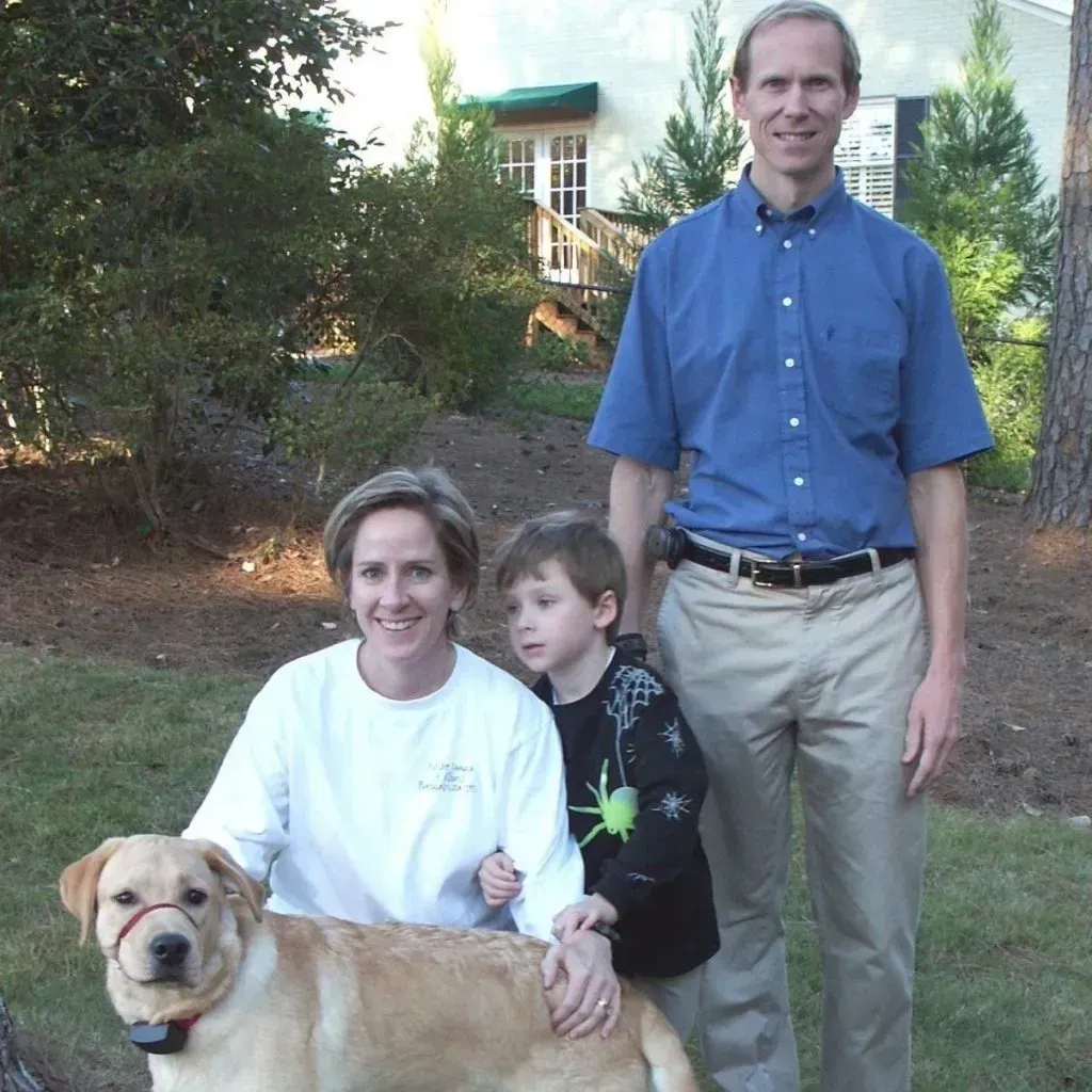 Family with dog poses outdoors; man in blue shirt, woman, child, yellow lab.