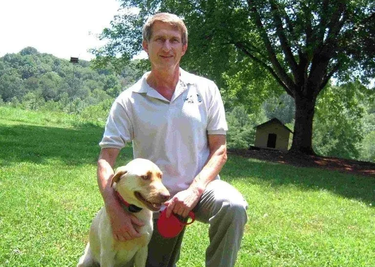 Man kneeling with a yellow Labrador on grass, dog house and trees in the background.