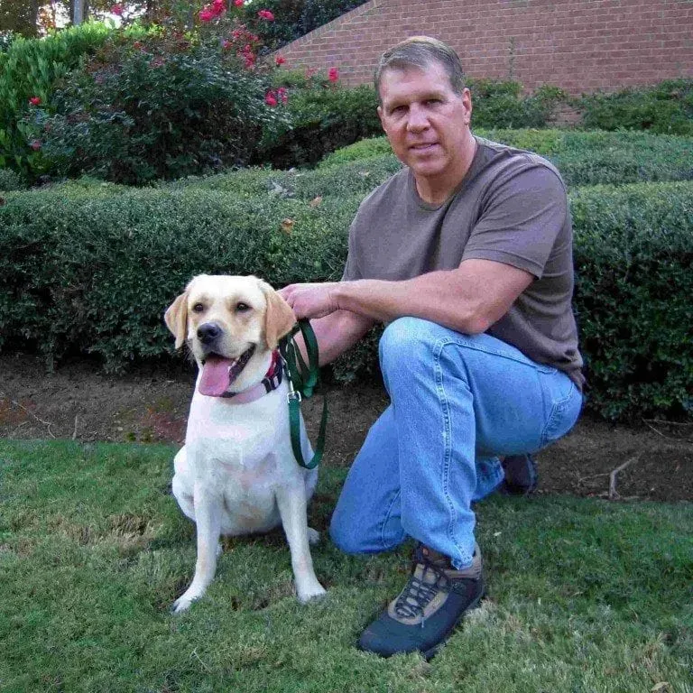 Man kneels with yellow Labrador on grass. Man wears blue jeans and a brown shirt. Bushes in background.
