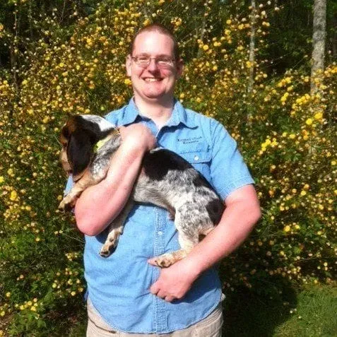 Man in glasses holding a blue tick hound, both smiling, in front of yellow flowering bushes.