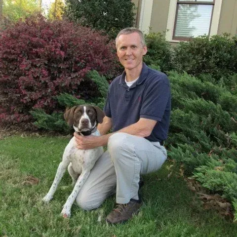 Man kneels with a dog in front of green bushes. The dog is spotted, and they are both smiling.