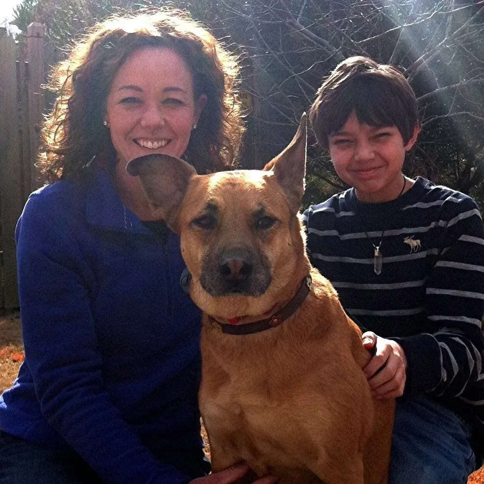Woman and boy smile with a tan dog outdoors. The dog has its ears perked up, and the boy has his arm around it.