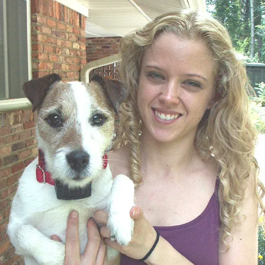 Woman with curly blonde hair smiles, holding a Jack Russell Terrier dog wearing a red collar outdoors.
