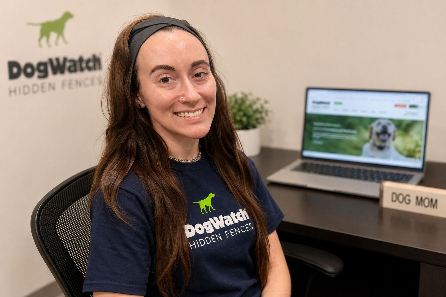 Woman in blue sweatshirt smiles at desk with laptop and monitor displaying website.