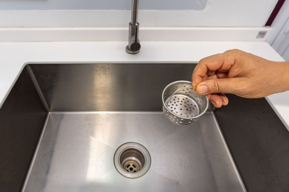 Hand holding a small, perforated metal strainer above a stainless steel kitchen sink drain.