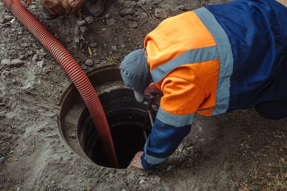 Man in workwear leans into a sewer. A red hose extends into the hole.