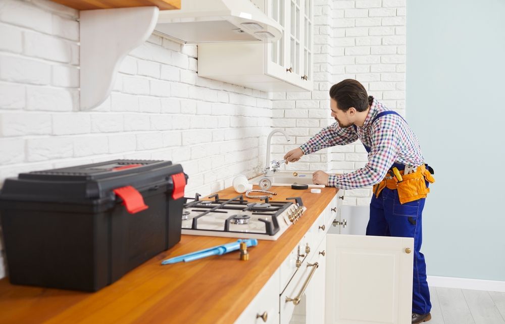 Plumber installing a kitchen faucet. He's wearing blue overalls and a tool belt, working in a white-walled kitchen.