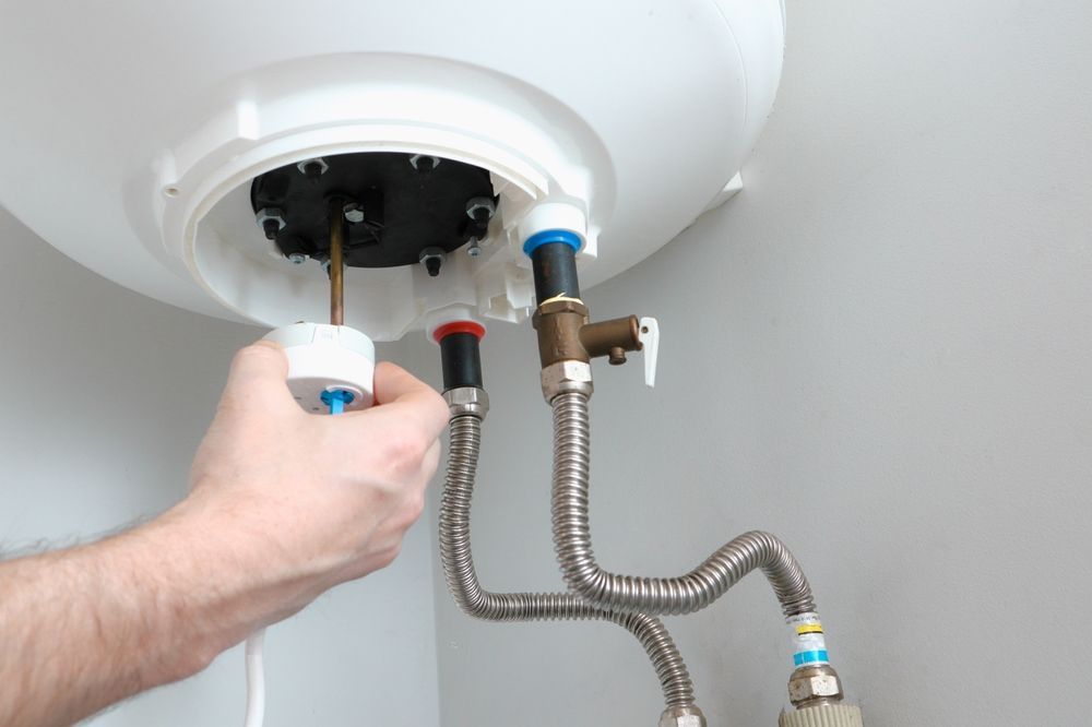 Hand installing a component on the top of a white water heater. Silver corrugated pipes connect to the heater.