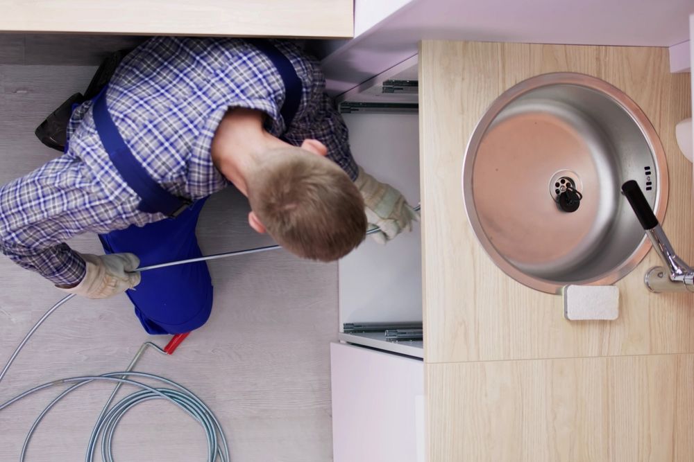 Plumber using a snake tool under a kitchen sink. The sink is stainless steel.