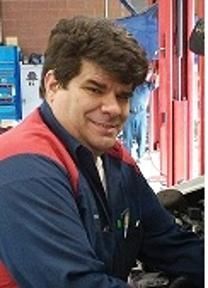 Man smiling in a blue and red work jacket in a shop or garage setting.